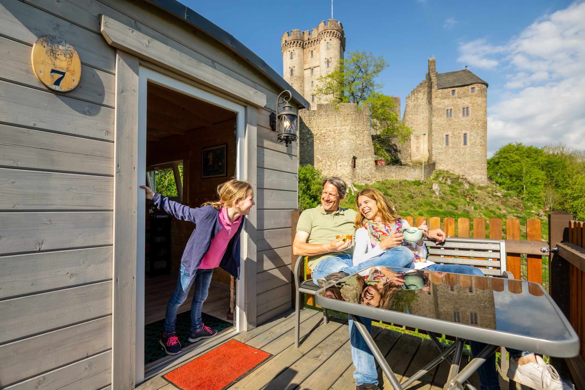 Eine Familie sitzt entspannt auf einer Terrasse vor einem kleinen Holzhaus. Im Hintergrund ist eine alte Burg und blauer Himmel zu sehen.