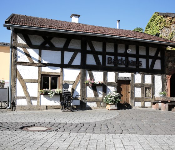 Half-timbered house with the inscription 'Backhaus', flowers in the windows, paved square, blue sky., © Historische Wassermühle Birgel, Mariette Spohr GmbH