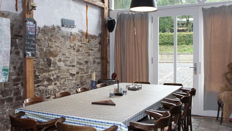 Een gezellige eetzaal met een lange tafel en houten stoelen. Grote ramen laten veel licht binnen en bieden uitzicht op de buitenlucht.