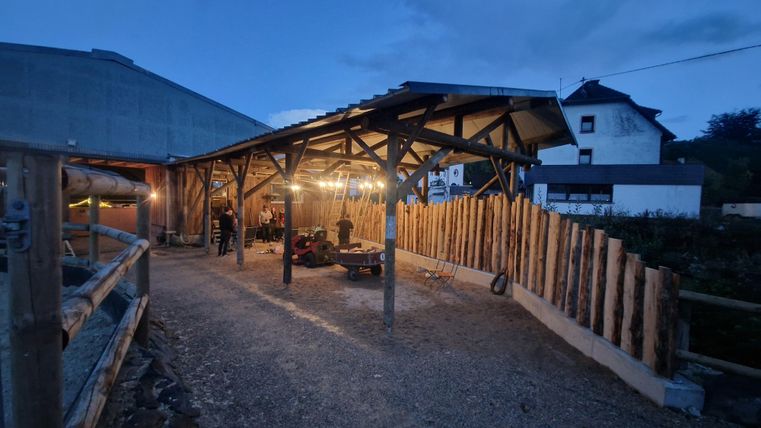 Une grange rurale à la tombée de la nuit avec un abri éclairé. À l'arrière-plan, on aperçoit des bâtiments et des troncs de bois.