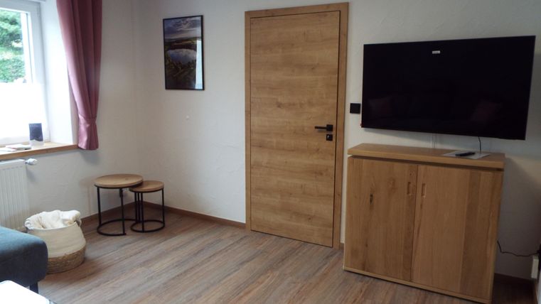 Ein modernes Wohnzimmer mit einem Fernseher an der Wand. Es gibt Holz- und Stoffmöbel sowie eine große Fenster mit Vorhängen.