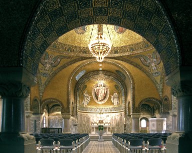 The interior of the Church of the Saviour with a view of the altar. The ceiling is decorated with golden mosaics, angels and a large chandelier. Numerous chairs form rows of seats for visitors.