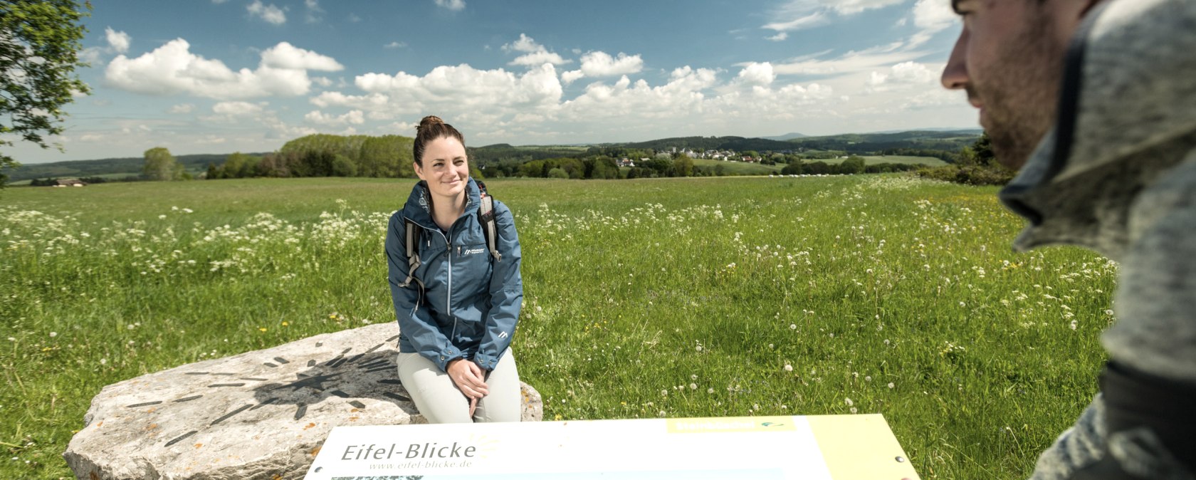 Eine Frau sitzt auf einem Stein, auf dem Richtungspfeile angebracht sind. Der Stein steht auf einer großen Wiesen, hinter der ein Dorf liegt. Im Vordergrund ist ein Mann, der an einer Informationstafel zu den Eifel-Blicken steht und die Frau anblickt.