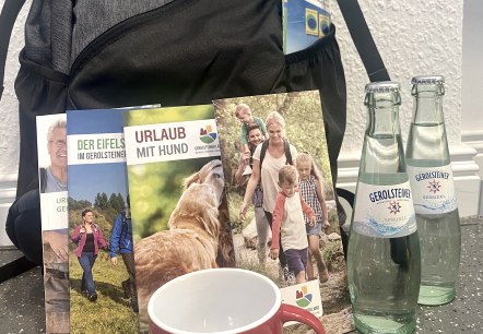 An Eifel rucksack with several tourist brochures, two Eifel mugs and two glass bottles of Gerolsteiner sparkling water.