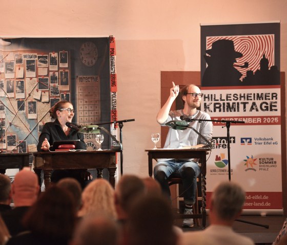 Zwei M&auml;nner und eine Frau sitzen nebeneinander an Tischen und lachen. Vor Ihnen dichtes Publikum und hinter Ihnen eine Indizienwand.