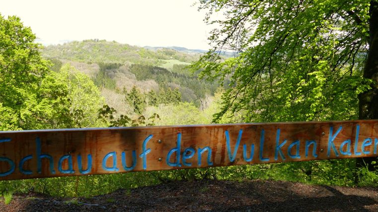 Un panneau en bois avec l'inscription « Regarde le volcan » se dresse dans un paysage forestier verdoyant. À l'arrière-plan, on peut voir des collines douces et un ciel dégagé.