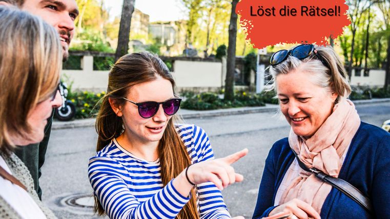 Eine Gruppe von vier Personen diskutiert und lächelt, während sie auf eine Karte schauen. Eine Person zeigt mit dem Finger auf einen Punkt und es gibt einen Text „Löst die Rätsel!“ im Bild.