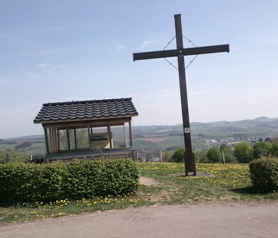 A large wooden cross and a small shelter stand on a hill overlooking a vast landscape under a blue sky., &copy; Touristik GmbH Gerolsteiner Land
