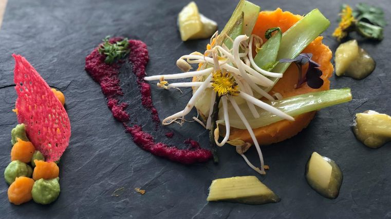 Un plat artistiquement présenté sur une assiette noire. Il contient différentes sortes de légumes, des sauces et des fleurs comestibles.