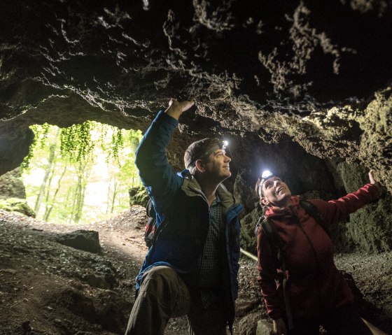 Zwei Personen mit Stirnlampen erkunden eine Höhle. Im Hintergrund sind Bäume und Tageslicht zu sehen., © Eifel Tourismus GmbH, Dominik Ketz