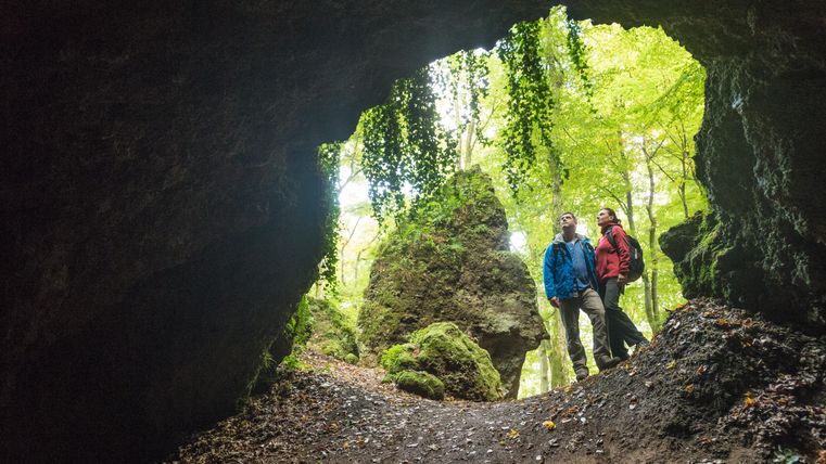 Zwei Wanderer stehen am Höhleneingang und schauen ins Innere. Hinter ihnen ein Wald mit hohen Bäumen und Felsen.