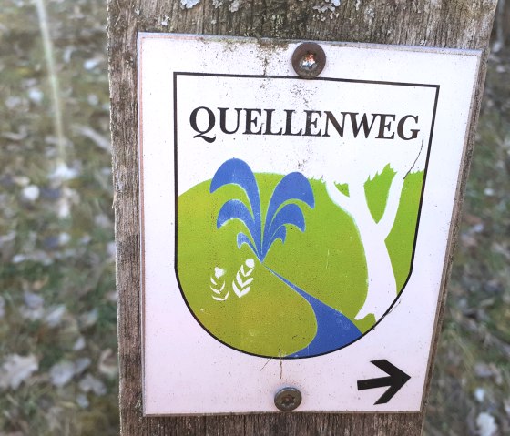Bord 'Quellenweg' met boom en waterloop op een groene achtergrond, bevestigd aan een houten paal., © Touristik GmbH Gerolsteiner Land, Ute Klinkhammer