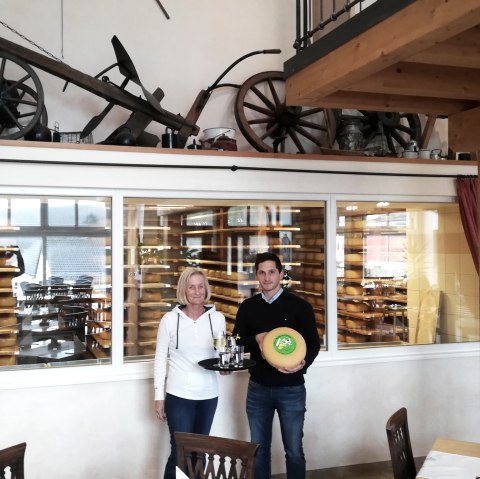 Ein Mann und eine Frau stehen vor eine gro&szlig;en Glasscheibe, hinter der in Regalen zahlreiche K&auml;selaibe reifen. Dei Frau tr&auml;gt ein Tablett mit Getr&auml;nken und der Mann einen K&auml;selaib.