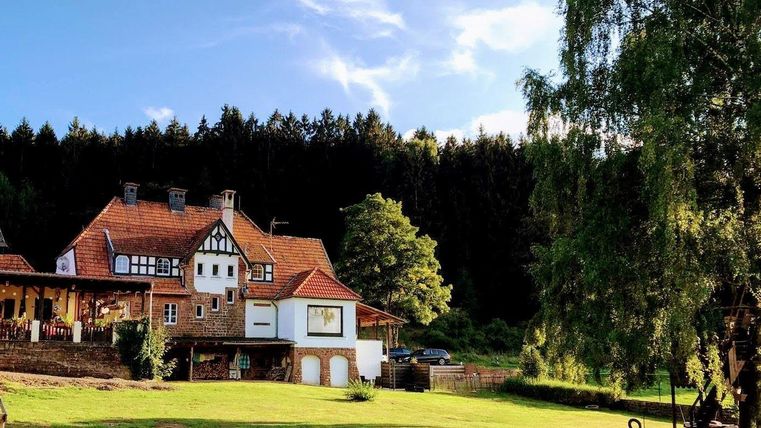 Une maison pittoresque avec une architecture traditionnelle, entourée d'un jardin verdoyant. À l'arrière-plan s'étend une forêt sous un ciel clair.