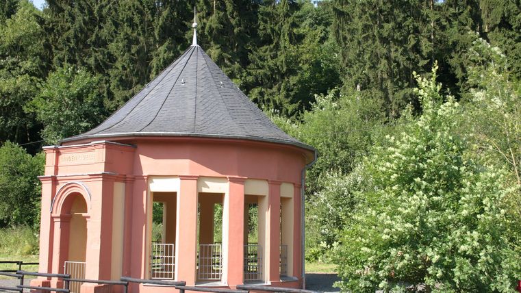 Ein elegantes Pavillon mit einem runden Dach und Säulen steht in einer grünen Umgebung. Die Bäume im Hintergrund verleihen der Szenerie eine ruhige Atmosphäre.