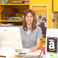Eine Frau steht an einem Postschalter und lächelt in die Kamera. Vor ihr ein Bildschirm mit Tastaur und hinter ihr Regale voller Umschläge, Pakete und weiterer Postartikel.