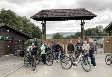 Negen sportief geklede personen staan voor de ingang van het Adler- en Wolfspark Kasselburg en enkele fietsen staan bij hen geparkeerd.