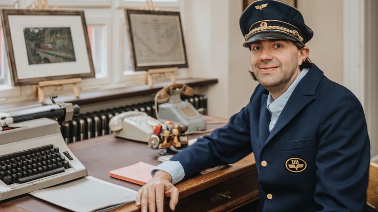 <p>Ein Mann in Uniform sitzt in einem Büro neben einem Schreibtisch mit Schreibmaschine, Telefon und Dokumenten. Große Fenster im Hintergrund.</p>