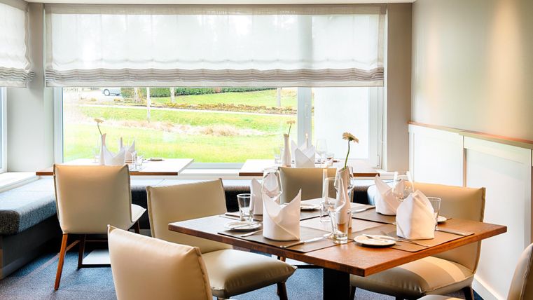 Elegantes Restaurant mit gedeckten Tischen, weißen Servietten und Blick auf grüne Landschaft durch großes Fenster.