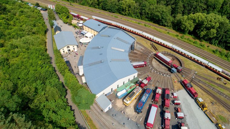 Eine Luftaufnahme eines Bahnbetriebswerks mit mehreren Lokomotiven und Waggons. Umgeben von Bäumen und Gleisen zeigt die Szene eine gut organisierte Eisenbahninfrastruktur.