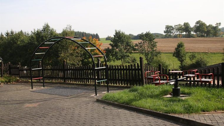 Ein Spielplatz mit einer Klettergerüst und einem eingezäunten Bereich. Im Hintergrund sind Wiesen und Bäume zu sehen.