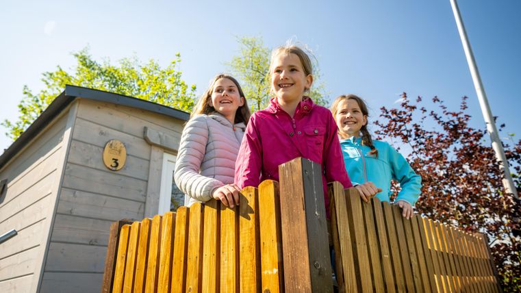 Drei fröhliche Mädchen stehen an einem Holzzaun und lächeln in die Kamera. Im Hintergrund sieht man einen kleinen Holzbau und grünes Laub.