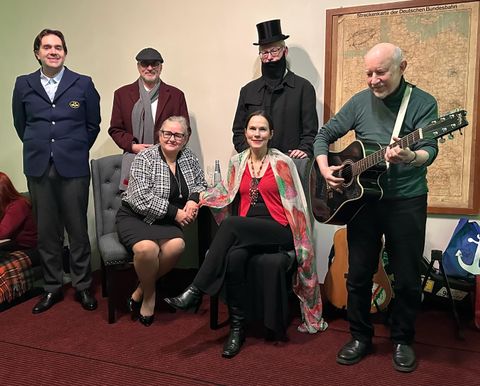 Sechs Personen versammeln sich in einem Raum. Drei sitzen, während die anderen drei stehen, und einer spielt Gitarre.