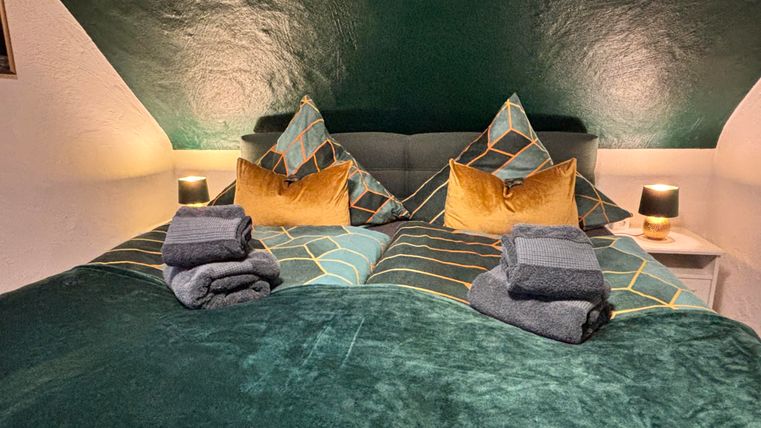 Ein gemütliches Schlafzimmer mit einem großen Bett, das mit grünen und goldenen Kissen dekoriert ist. Neben dem Bett stehen zwei kleine Lampen und frisch gefaltete Handtücher.