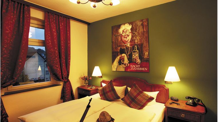 Une chambre confortable avec deux lits, des coussins décorés et une grande fresque murale. Les fenêtres sont ornées de rideaux rouges et il y a deux tables de nuit avec des lampes.