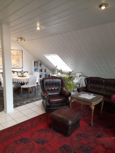 Ein gemütliches Wohnzimmer mit braunen Ledersesseln und einem kleinen Tisch. Im Hintergrund sind ein Esstisch und Regale sichtbar, unter einem schrägen Dachfenster.