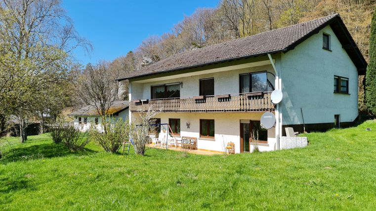 A modern house surrounded by green lawn and trees. The facade is bright, and there is a balcony on the upper floor.
