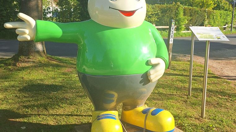 Eine bunte Statue eines lächelnden Charakters in einem grünen Shirt und einer Mütze. Der Charakter steht auf einer Wiese und zeigt mit dem Finger nach vorne.