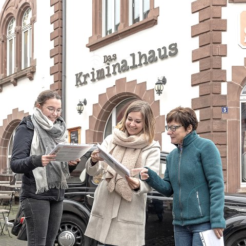 Drei Frauen stehen lachend vor dem Gebäude „Das Kriminalhaus“ und halten Unterlagen in der Hand.  Im Hintergrund parkt ein schwarzes Auto vor dem historischen Haus mit roten Sandsteinrahmen.