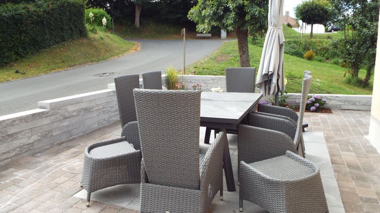 Ein gemütlicher Außenbereich mit einem Tisch und acht Stühlen aus grauem Rattan. Die Terrasse ist umgeben von Pflanzen und einem ruhigen Weg.