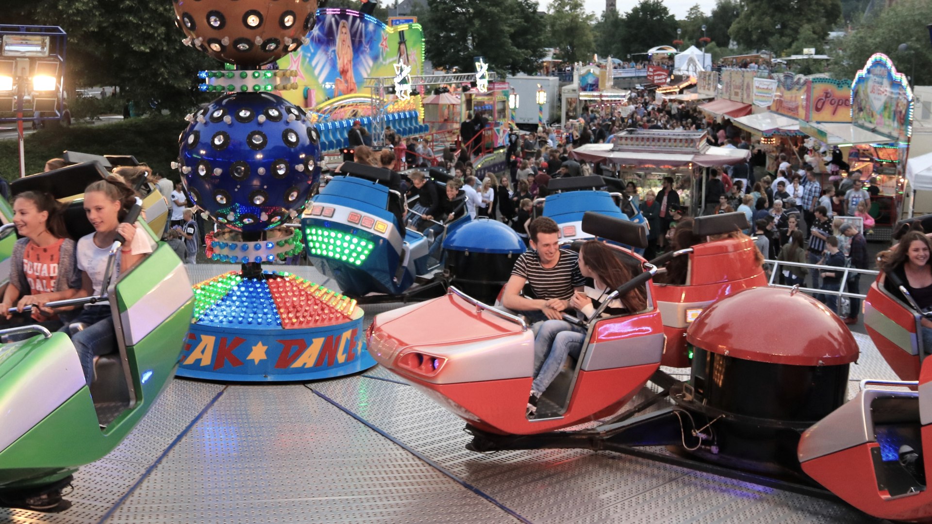 BreakDancer ride with passengers. Behind it, colourful fairground hustle and bustle with lots of visitors and showmen.