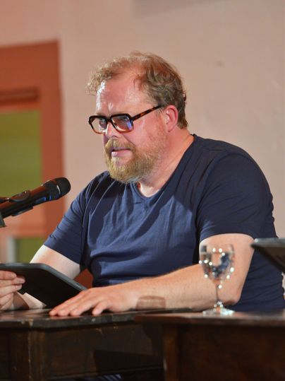 Ein man sitzt an einem Tisch und liest von einem Tablett in ein Mikrophon vor.