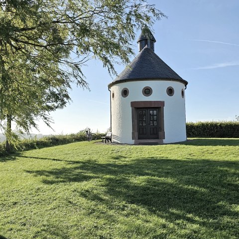 Ronde kapel met een zwart dak op een groene weide, omringd door bomen en een blauwe lucht. Een fiets leunt tegen de kapel., © Touristik GmbH Gerolsteiner Land