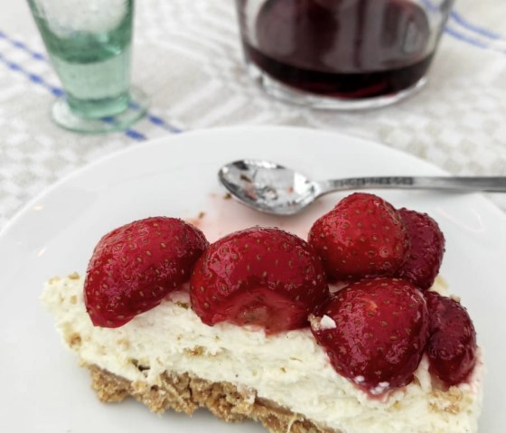 Ein St&uuml;ck Erdbeerkuchen auf einem Teller, daneben ein Glas und eine Karaffe mit rotem Getr&auml;nk auf einem Tisch., &copy; Touristik GmbH Gerolsteiner Land, Ute Klinkhammer