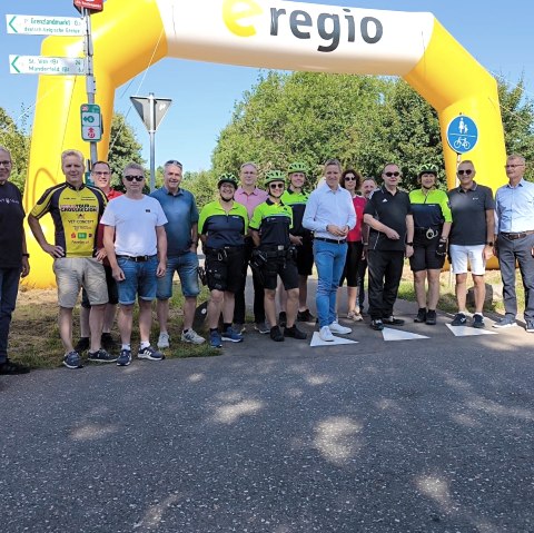 Zahlreiche Personen, welche teilsweise sportlicher Radfahrkleidung tragen, stehen auf dem Radweg unter einem gro&szlig;en Luftbogen des Sponsors e-regio.