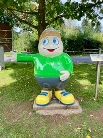 Statue von Maskottchen Willi Basalt mit grünem Shirt, grauer Hose und gelben Schuhen zeigt nach links. Hinter ihm ein Baum und ein Park.