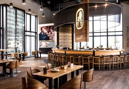 Restaurant area of the Bitburger Brewery with many tables, chairs and a round bar in the centre. Large window walls and matching decoration.