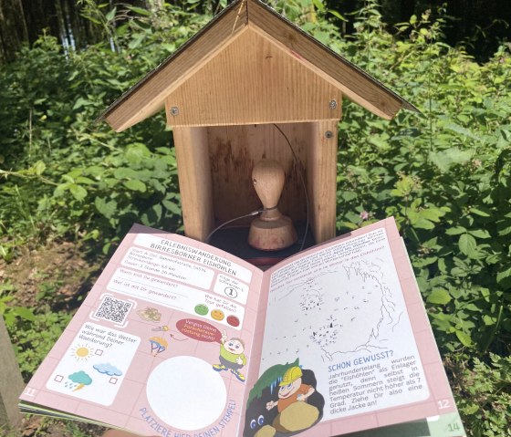Ein Wanderpass mit Informationen zur Erlebniswanderung Birresborner Eishöhlen liegt offen vor einem Holzstempelhäuschen im Wald., © Touristik GmbH Gerolsteiner Land, Leonie Post