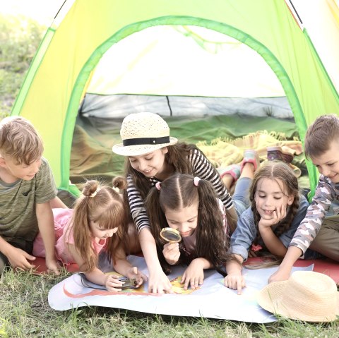 Zes kinderen liggen en zitten dicht tegen elkaar aan voor een kleurrijke tent op een weiland en bekijken een kaart die voor hen ligt. Sommige kinderen wijzen naar de kaart en één kind bekijkt de kaart nauwkeuriger met een vergrootglas.