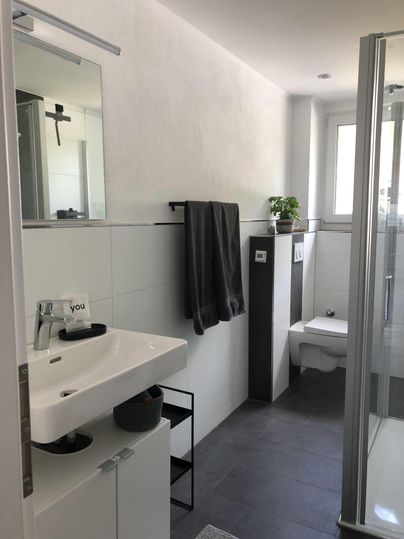 Ein modernes Badezimmer mit einer Dusche und einem Waschbecken. Es sind helle Wände, dunkle Fliesen und ein Pflanzenarrangement zu sehen.