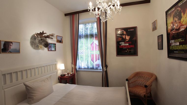 Une chambre confortable avec un lit, un chandelier et des affiches de films décoratives sur le mur. La fenêtre laisse entrer la lumière du jour, créant une atmosphère accueillante.