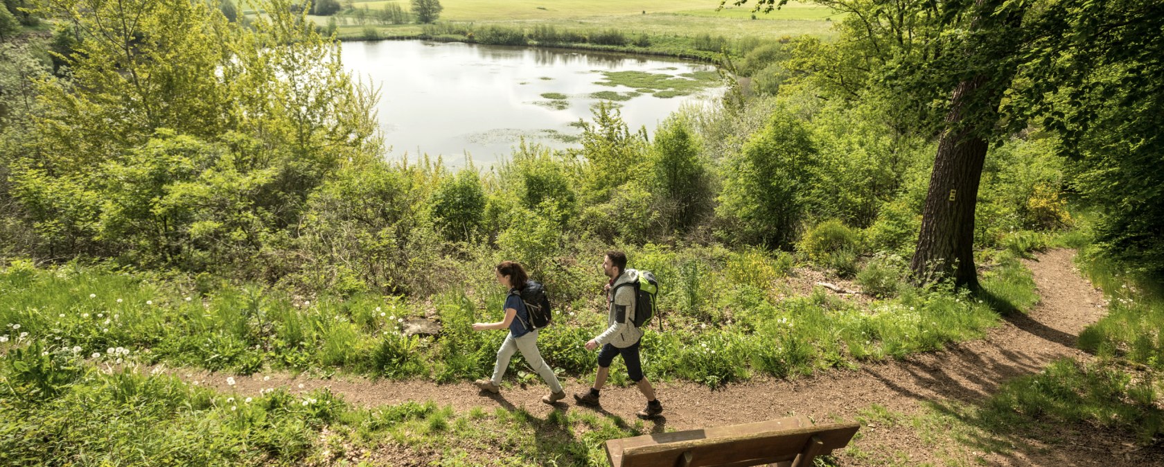 Zwei Wanderer auf einem Pfad am Eichholzmaar in der Eifel. Im Hintergrund der Maarsee und grüne Landschaft. Eine Bank steht am Wegesrand., © Eifel Tourismus GmbH, Dominik Ketz
