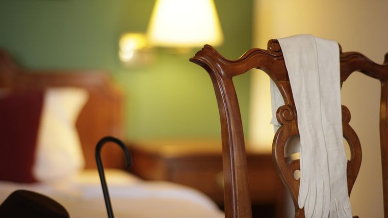Un extrait stylé de chambre d'hôtel avec une chaise en bois sur laquelle reposent des gants blancs. À l'arrière-plan, on aperçoit un lit et une lampe.