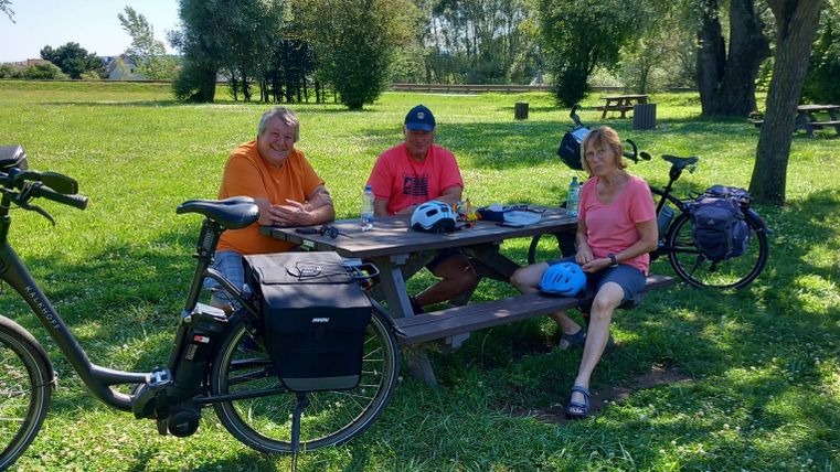 Drei Personen sitzen an einem Tisch im Freien und entspannen sich. Im Hintergrund sind Fahrräder und eine grüne Wiese zu sehen.
