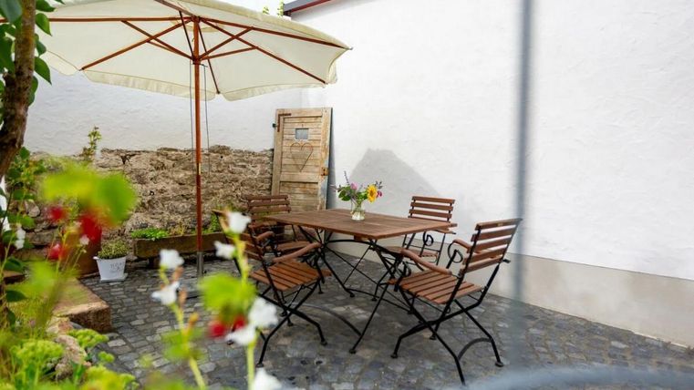 Ein schöner Außenbereich mit einem Tisch und Stühlen unter einem Sonnenschirm. Umgeben von einer blühenden Pflanzenlandschaft und einer weißen Wand.