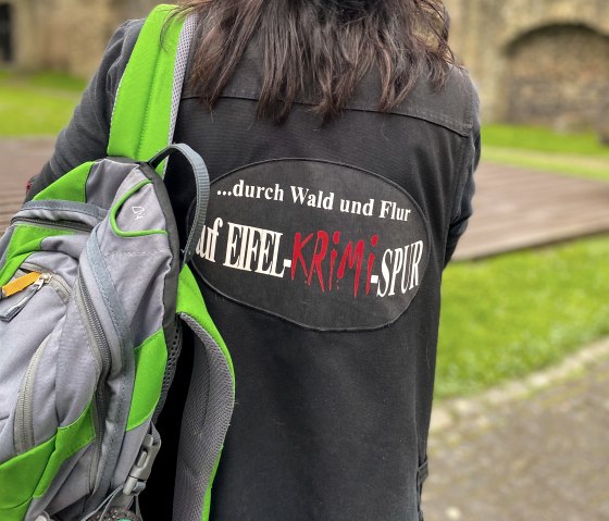 Eine Hut tragende Person steht mit dem R&uuml;cken zum Betrachter vor einer gro&szlig;en Stadtmauer. Sie tr&auml;gt eine Jacke mit der Aufschrift "...durch Wald und Flur auf Eifel-Krimi-Spur" auf dem R&uuml;cken und tr&auml;gt &uuml;ber einen Schulter einen Rucksack.